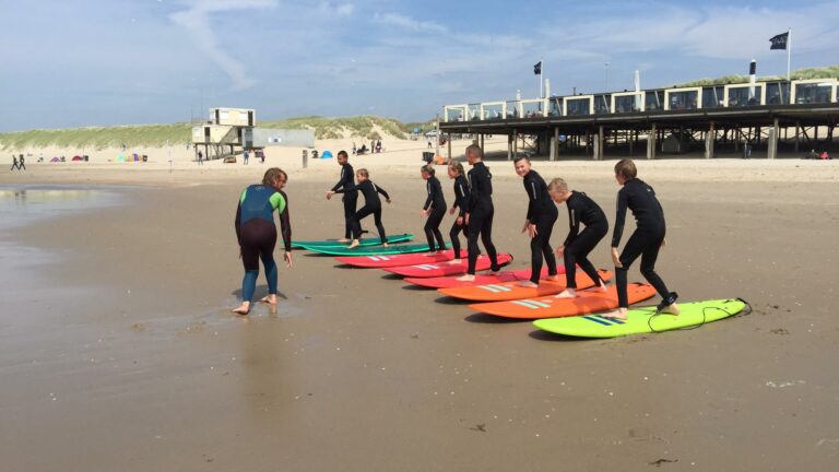 Kinderen leren surfschool op het strand bij Surfschool High 5 in Julianadorp met op de achtergrond restaurant Paal 6